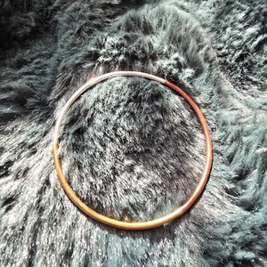 May include: A two-tone metal bracelet, with a silver and copper finish, lies on a textured, dark teal surface. The bracelet is a simple, circular design, showcasing a minimalist aesthetic. The contrast between the metals and the background creates a visually appealing piece of jewelry.