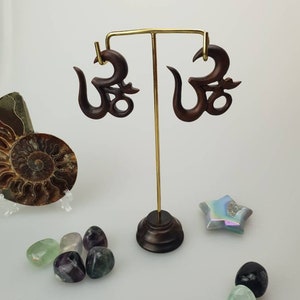 Può includere: Orecchini in legno marrone scuro a forma di simbolo Om, esposti su un supporto dorato. Gli orecchini sono circondati da pietre preziose lucide e una stella decorativa. Il simbolo Om è un'icona spirituale.