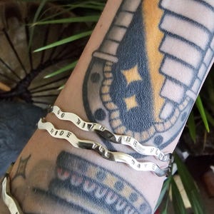 May include: Three silver-colored, wavy bracelets with the words "GO WITH THE FLOW RIDE THE WAVE" engraved on them. The bracelets are worn on an arm with tattoos. The bracelets are made of a shiny metal.
