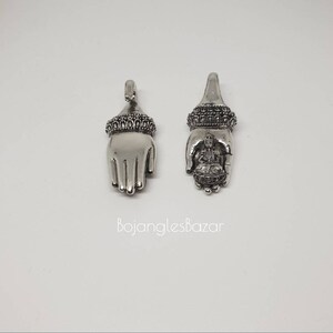 May include: Two silver-toned hand-shaped pendants. One pendant features an open hand design, while the other holds a small figure within the palm. Both have ornate detailing at the wrist and a loop for hanging. The text "BojanglesBazar" is at the bottom.