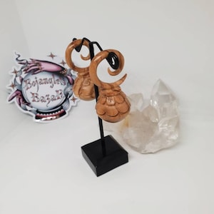 May include: A pair of light brown wooden ear gauges featuring a spiral top and a rounded, segmented bottom section. The gauges are displayed on a black metal stand. A sticker with the text Bojangles Bazaar and a crystal are visible nearby.