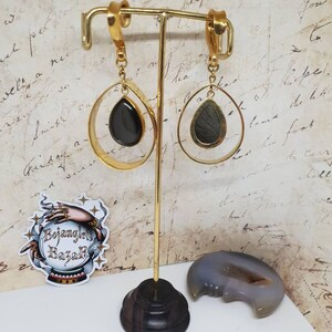 May include: Gold-toned earrings with a teardrop-shaped, dark stone pendant. The earrings are displayed on a gold-colored stand with a dark wooden base. A sticker with the text "Bojangles Bazar" is visible.