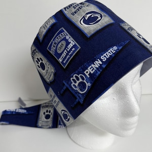 Peut inclure: Un bonnet de gommage bleu marine de l'Université Penn State. Le bonnet présente des motifs de fanions et d'écussons avec les mots "Penn State", "Nittany Lions" et "University Park PA" en blanc. Le bonnet est sur une tête de mannequin blanche.