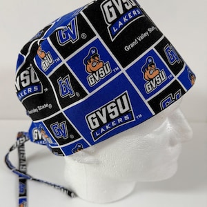 Peut inclure: Un bonnet de chirurgien à motifs bleus, noirs et blancs, avec le texte "GVSU Lakers" et une mascotte de dessin animé. Le bonnet est conçu avec un motif quadrillé, et les liens sont visibles sur le côté. Les mots "Grand Valley State" sont également visibles.