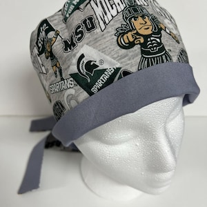 Peut inclure: Un bonnet de chirurgien à motifs gris et verts avec une cravate grise. Le bonnet présente le logo et la mascotte de l'Université d'État du Michigan.