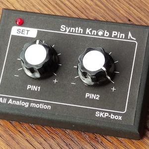 Könnte beinhalten: Eine schwarze Box mit zwei Knöpfen, die mit "PIN1" und "PIN2" beschriftet sind. Die Box hat eine rote LED mit der Aufschrift "SET" sowie den Text "Synth Knob Pin" und "SKP-box". Die Box befindet sich auf einer Holzoberfläche.