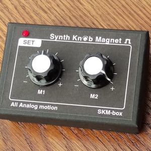 May include: Black box with two knobs labelled "M1" and "M2" with plus and minus signs. The box has a red LED labelled "SET" and the text "Synth Knob Magnet ♫ All Analog motion SKM-box".
