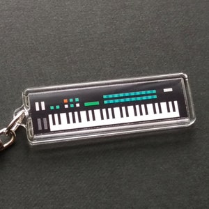 May include: A clear keychain featuring a black and white image of a synthesizer with a teal and green control panel. The keychain has a silver metal ring.