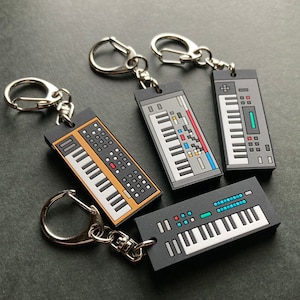 Op de afbeelding: Vier sleutelhangers met verschillende vintage-stijl synthesizers. Elke sleutelhanger heeft een zilveren sleutelring en een zwart-wit ontwerp met kleurrijke accenten.