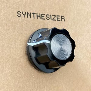 May include: A black synthesizer knob with a silver, circular design, set against a tan backdrop. The word "SYNTHESIZER" is printed above the knob in black, block letters. The knob has a ridged edge.