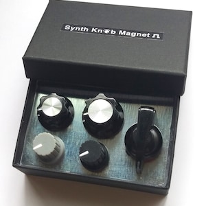Op de afbeelding: Een zwarte doos met de tekst "Synth Knob Magnet П" op de deksel. De doos bevat vijf knoppen, vier ronde en één met een puntige bovenkant. Twee van de ronde knoppen zijn zwart met een zilveren midden, één is grijs en één is zwart.