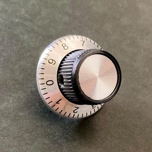 May include: A silver-colored rotary knob with a black center and a circular design. The knob has numbers from 0 to 9 around the edge. The center of the knob has a textured, circular pattern.