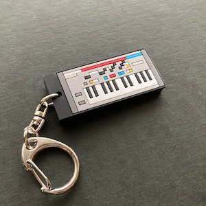 May include: A black and gray keychain shaped like a vintage synthesizer with a silver key ring. The synthesizer has a white keyboard, black and white keys, and colorful buttons.