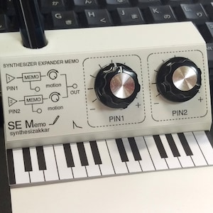 May include: A white synthesizer expander module with two black knobs labelled "PIN1" and "PIN2". The module has a diagram with the text "MEMO" and "motion" and a keyboard below it.