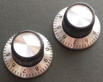 Puede incluir: Dos perillas plateadas y negras con los números del 4 al 9 impresos alrededor del borde. Las perillas tienen una parte superior de metal plateado y una base de plástico negro.