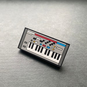 May include: A black and gray enamel pin shaped like a vintage synthesizer with a white keyboard, red, blue, and yellow buttons, and a black border.