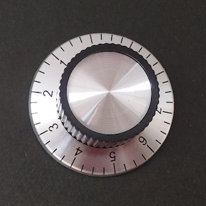 Könnte beinhalten: Ein silberfarbener Drehknopf mit einem schwarzen Gummi-Griff und einem nummerierten Zifferblatt von 1 bis 9. Der Knopf hat ein kreisförmiges Design mit geriffelter Kante und einem glatten, reflektierenden Zentrum. Die Zahlen sind in einer einfachen, gut lesbaren Schrift.