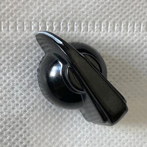 Könnte beinhalten: Ein glänzender schwarzer Knopf mit rundem Sockel und einem flachen, abgewinkelten Griff. Der Knopf hat eine glatte, reflektierende Oberfläche, mit einem kleinen Bereich weißer Farbe am Rand des Griffs. Wahrscheinlich ein Möbel- oder Gerätebauteil.