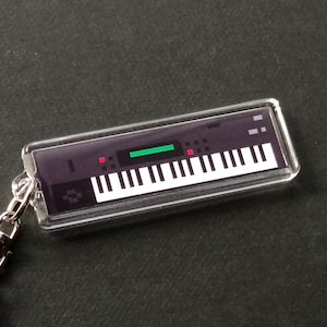 May include: A clear keychain featuring a black and white keyboard design. The keyboard has a green display and red buttons.
