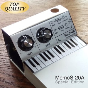 May include: A silver and black MemoS-20A Special Edition synthesizer notepad holder with a small keyboard and two knobs. The notepad holder has a silver metal clip and a white notepad.