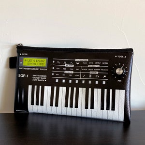 May include: Black zippered pouch with a synthesizer design. The pouch has a white keyboard, black knobs, and the text "Let's Enjoy Synth Life!" and "Synthesizer Gadget Pouch".