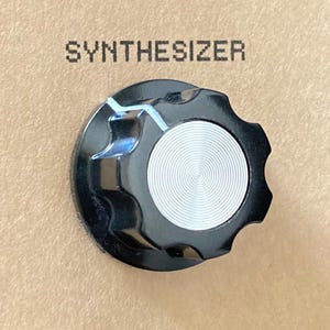 Könnte beinhalten: Nahaufnahme eines schwarzen Synthesizer-Knopfes mit silbernem Mittelteil und geriffelten Kanten. Das Wort "SYNTHESIZER" ist über dem Knopf aufgedruckt. Der Knopf befindet sich vor einem neutralen Hintergrund.