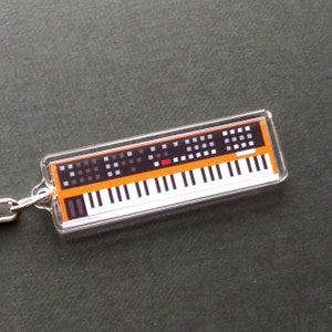 Könnte beinhalten: Ein durchsichtiger Acryl-Schlüsselanhänger mit einem orangefarbenen und schwarzen Synthesizer im Vintage-Stil mit einer Tastatur und Drehknöpfen.