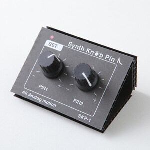 May include: A black and grey cardboard synthesizer with two knobs labelled "PIN1" and "PIN2". The text "Synth Knob Pin" and "SKP-1" are printed on the front of the synthesizer.