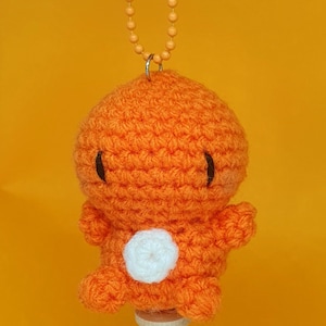 May include: Orange crocheted plush toy with a white circle on its belly, hanging from a chain with a small orange bell.