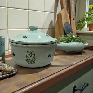 Op de afbeelding: Een lichtgroene keramische ovenschaal met deksel en handvat. De schaal is versierd met een tulpontwerp en kleine bladaccenten. De ovenschaal staat op een houten aanrecht in een keuken.