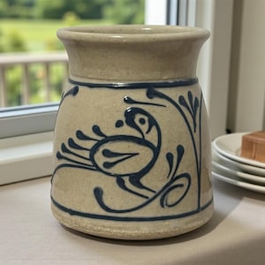 Könnte beinhalten: Ein beigefarbener Keramiktopf mit ausgestelltem Rand und rundem Boden. Der Topf ist mit einem blauen Vogel- und Blumendesign verziert. Der Topf ist etwa 13 cm hoch.