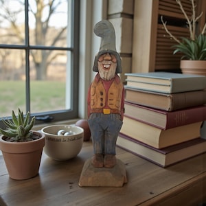 May include: A hand-carved wooden gnome figurine. The gnome wears a gray hat, a tan vest over a red shirt, and blue pants. The figurine stands on a small wooden base. The gnome has a white beard and a smiling expression.