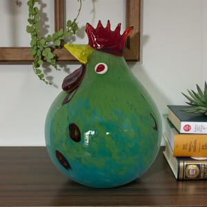 May include: A round, hand-blown glass sculpture shaped like a rooster. The body is a vibrant mix of green and teal glass with dark spots. It features a bright yellow beak, a deep red comb on top, and a dark red wattle under the chin.