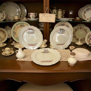 May include: A display of antique porcelain dishware, including plates, cups, and saucers, arranged on a dark wooden surface and within a glass-fronted cabinet. The plates feature a floral and teacup design, with scalloped edges. A small white creamer and salt shaker are also visible.