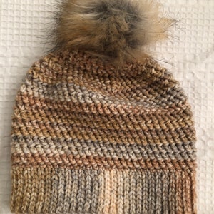 May include: A brown, beige, and cream knitted beanie with a fluffy brown pom pom.
