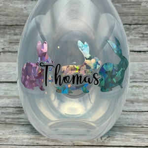 Könnte beinhalten: Klares Plastik-Osterei mit schimmernden Hasenaufklebern und dem Namen "Thomas" in Schwarz.