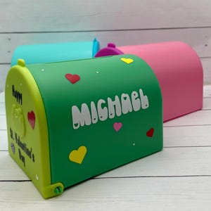 Op de afbeelding: Groene brievenbus met de naam Michael in witte letters. De brievenbus heeft een rood hart, een geel hart en een roze hart erop. De brievenbus zegt ook "Gelukkig Valentijnsdag".