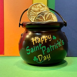 Può includere: Un calderone nero con manico, decorato con trifogli verdi e il testo "Happy Saint Patrick's Day". Monete d'oro con disegni di trifogli sono all'interno del calderone.