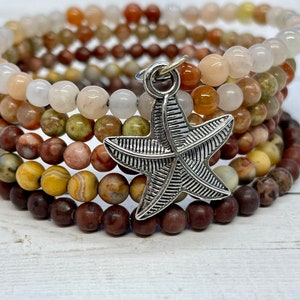 Puede incluir: Un brazalete de varias capas hecho de varias piedras marrones, beige y blancas con un dije de estrella de mar plateado.