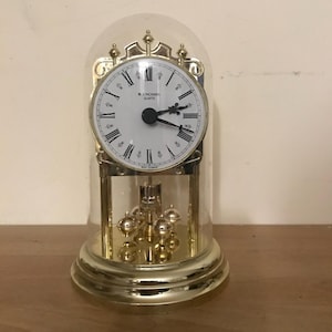 Può includere: Un orologio dorato con una cupola di vetro e un quadrante bianco. L'orologio ha un pendolo e numeri romani. Il quadrante dell'orologio indica "Junghans' Quartz".