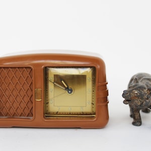 Puede incluir: Un radio despertador vintage marrón con una esfera dorada y agujas negras. El radio despertador está sobre una superficie blanca junto a una pequeña figura de oso marrón.