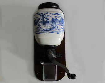 May include: A vintage, white ceramic coffee grinder with a blue and white Delftware design featuring a windmill and houses. The grinder has a wooden base and a metal crank.