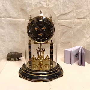 May include: An antique clock with a black face, gold accents, and a glass dome. The clock features floral designs and a pendulum. The base is dark blue with gold trim. A small bear figurine and a notepad are also present.