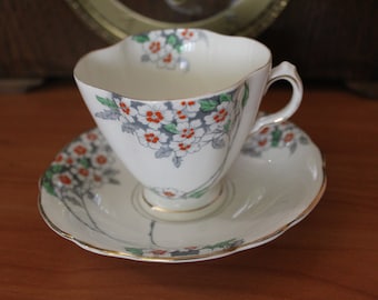 Puede incluir: Una taza de té y un platillo blancos con un patrón floral en tonos de blanco, rojo, verde y gris. El diseño floral presenta pequeñas flores blancas con centros rojos y hojas verdes.