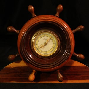 Puede incluir: Un reloj de madera con forma de rueda de barco con una esfera dorada y marcadores de hora romanos negros. El reloj está montado sobre una base de madera.