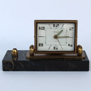 May include: A gold-toned alarm clock with a white face and black hands sits on a black marble base. The clock face has the words "Tambour" and "Paris" printed on it.
