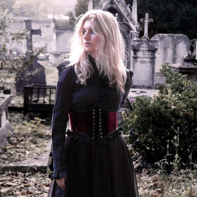 May include: A dark navy blue dress with a burgundy corset. The corset is laced up the front. The dress has long sleeves with ruffled cuffs. The background is a cemetery with stone monuments and greenery.