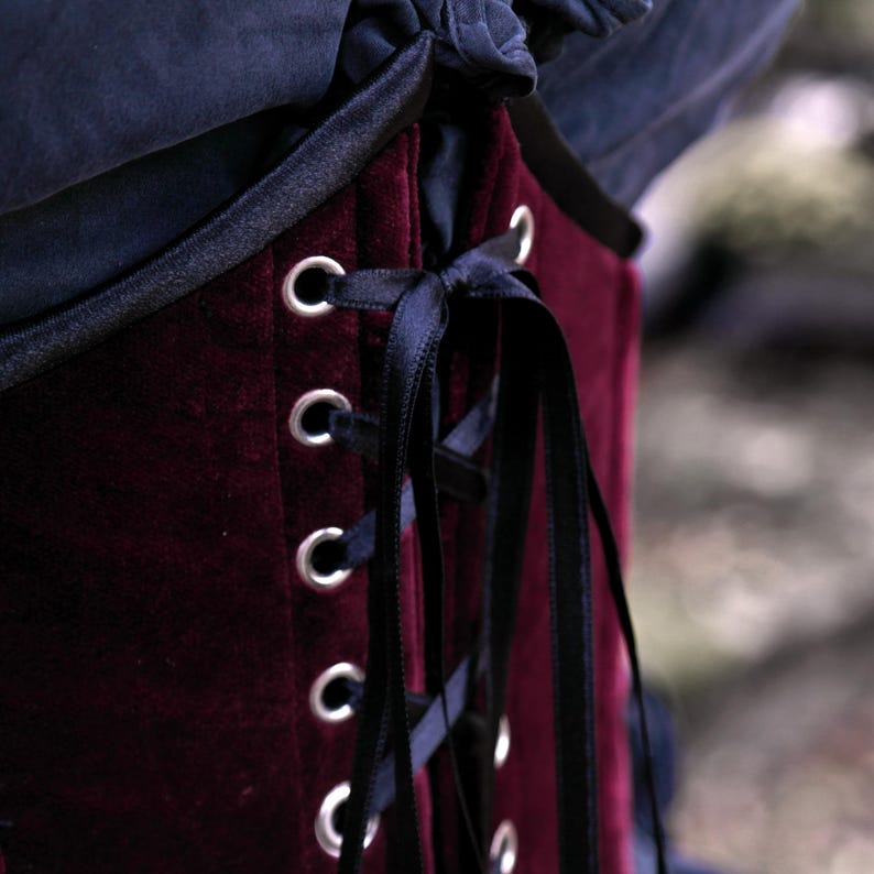 May include: Close-up of a burgundy velvet corset with black trim and lacing. The corset features silver eyelets and a black satin ribbon for closure. The garment is part of a gothic or historical fashion ensemble.