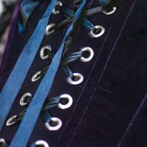 May include: Close-up of a dark purple velvet corset with silver eyelets and black and blue satin ribbons. The corset has vertical seams and a lace trim at the top edge. The ribbons are laced through the eyelets.