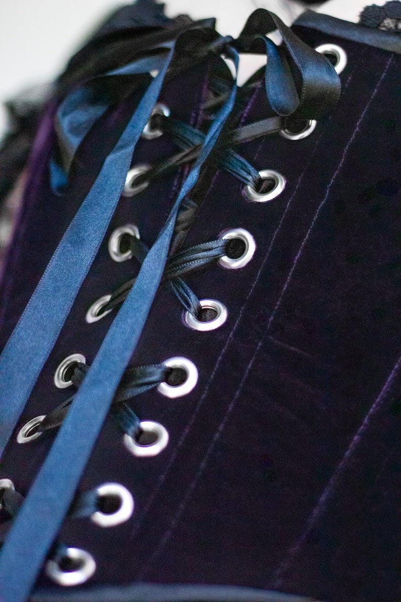May include: Close-up of a dark purple velvet corset with silver eyelets and black and blue satin ribbons. The corset has vertical seams and a lace trim at the top edge. The ribbons are laced through the eyelets.